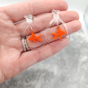 May include: A pair of clear acrylic earrings with a dangling orange goldfish charm. The earrings are shaped like a teardrop and have a silver hook.