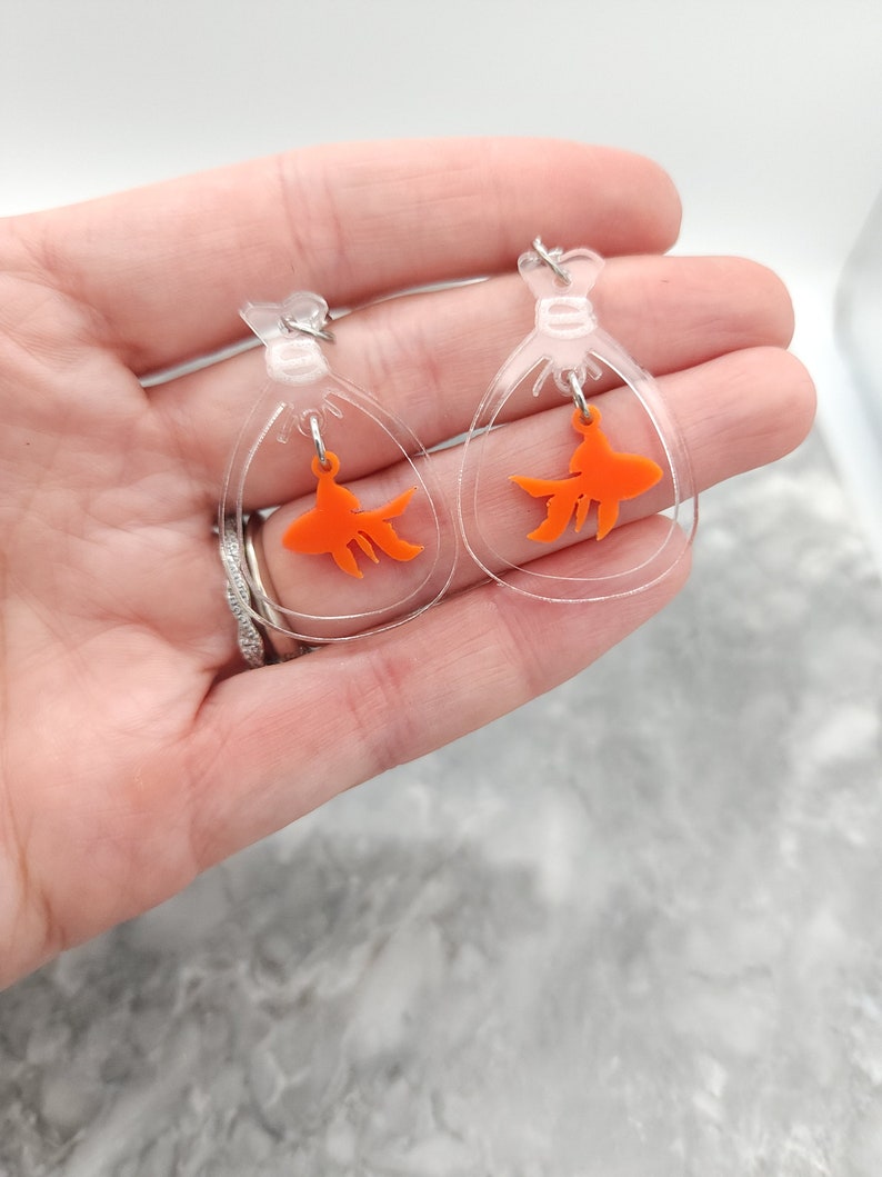 May include: A pair of clear acrylic earrings with a dangling orange goldfish charm. The earrings are shaped like a teardrop and have a silver hook.