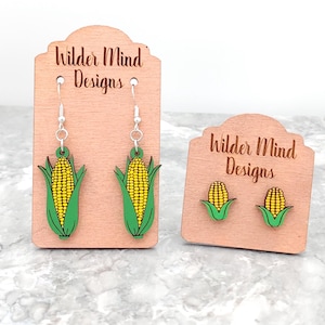 May include: Two pairs of corn on the cob earrings. The larger pair is dangle earrings with silver hooks. The smaller pair is stud earrings. Both pairs are made of wood and painted yellow, green, and white.