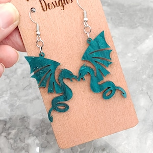 May include: Teal green dragon earrings with silver hooks. The dragons are facing each other and have a sparkly finish. The earrings are displayed on a wooden card with the text "Wilder Mind Designs".