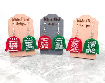 May include: Six pairs of Christmas sweater earrings. Each pair features a different red and green sweater design with white text. The text on the sweaters includes phrases like "Making a list and checking it thrice", "Best wishes warmest regards", "Its part of who you are", "Feats of strength airing of grievances", "Happy birthday Jesus", and "I'm dreaming of a Dwight Christmas".