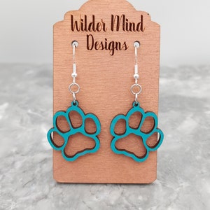 May include: Teal blue paw print earrings on a wooden display stand with the text "Wilder Mind Designs".