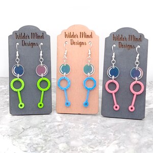 May include: Three pairs of dangle earrings with a bubble wand design. The earrings are on display cards with the text "Wilder Mind Designs". The bubble wands are green, blue, and pink.