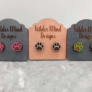 May include: Wooden paw print stud earrings in three color variations: pink and brown, natural wood, and green and brown. Each pair is displayed on a small wooden display card with the text "Wilder Mind Designs" engraved on it.