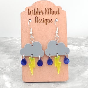 May include: Gray cloud earrings with yellow lightning bolts and blue raindrops hanging from them. The earrings are on a wooden display card with the text "Wilder Mind Designs".