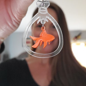 May include: Clear acrylic dangle earrings with a gold fish charm hanging inside. The fish is orange and the earrings are shaped like a bag.