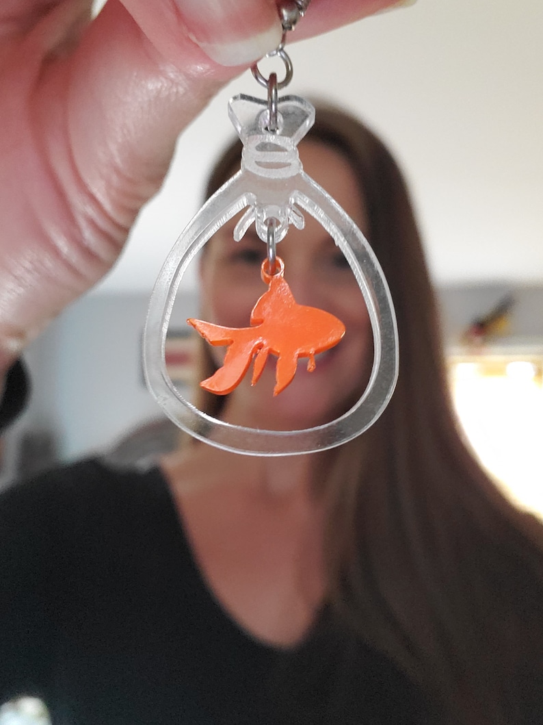 May include: Clear acrylic dangle earrings with a gold fish charm hanging inside. The fish is orange and the earrings are shaped like a bag.
