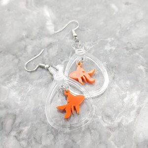 May include: A pair of clear acrylic earrings with a dangling orange goldfish charm. The earrings are shaped like a teardrop and have a silver hook.