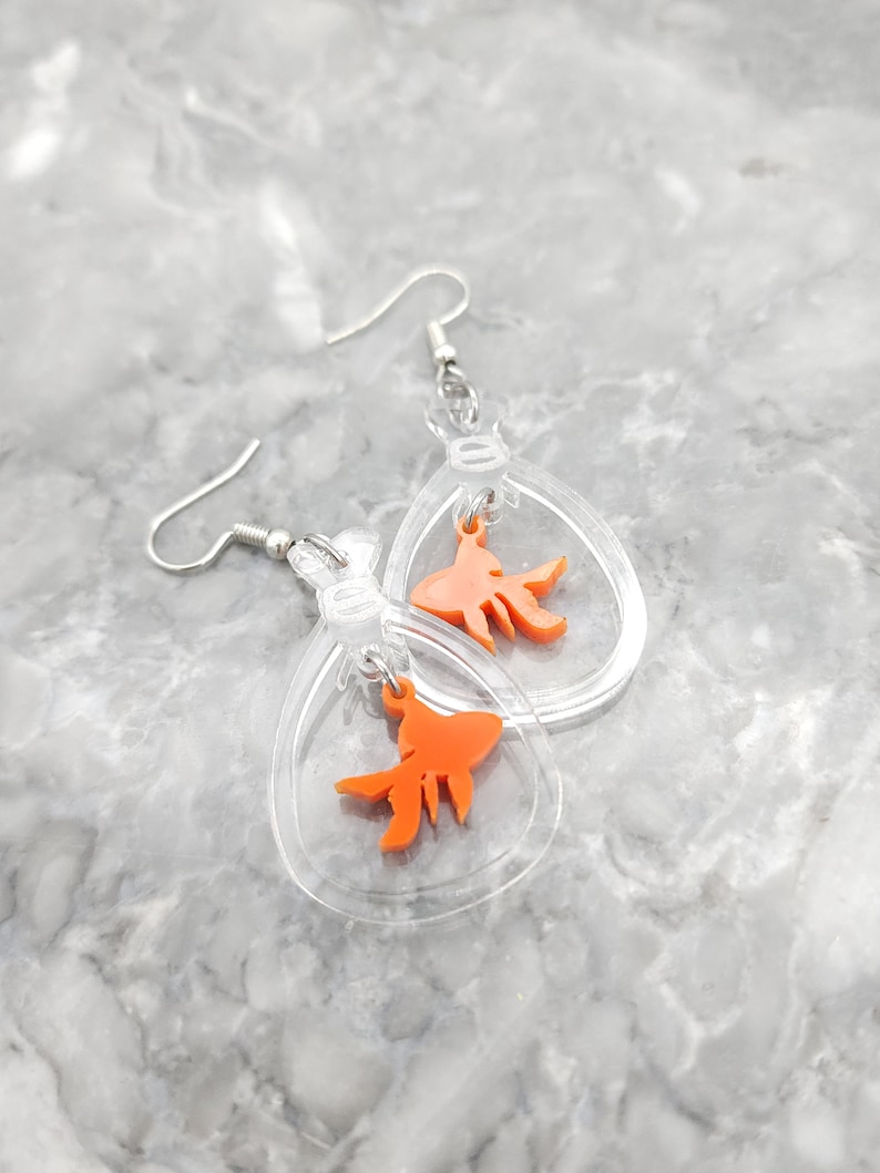 May include: A pair of clear acrylic earrings with a dangling orange goldfish charm. The earrings are shaped like a teardrop and have a silver hook.