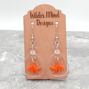 May include: Clear acrylic earrings with a gold fish inside each earring. The earrings are hanging from a wooden display card with the text "Wilder Mind Designs".