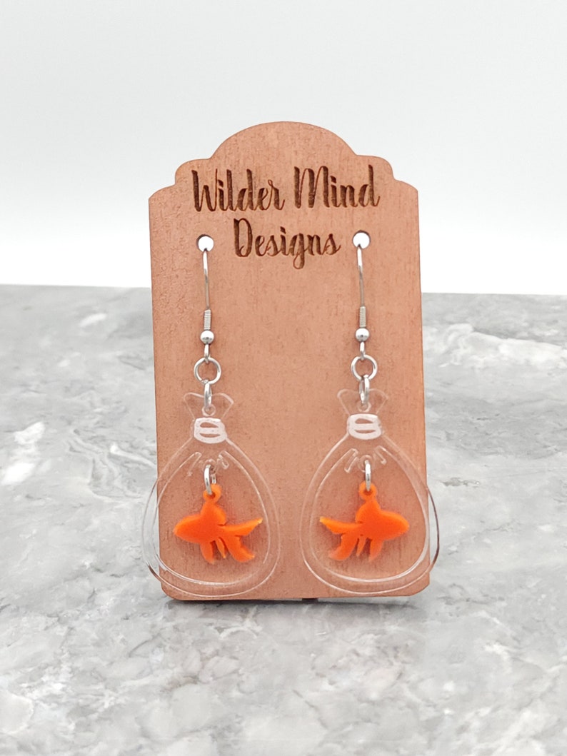 May include: Clear acrylic earrings with a gold fish inside each earring. The earrings are hanging from a wooden display card with the text "Wilder Mind Designs".