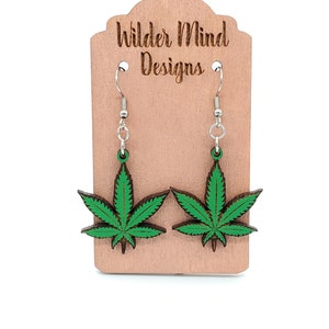 May include: A pair of green marijuana leaf earrings on a wooden display card with the text "Wilder Mind Designs".