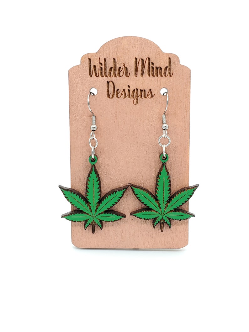 May include: A pair of green marijuana leaf earrings on a wooden display card with the text "Wilder Mind Designs".