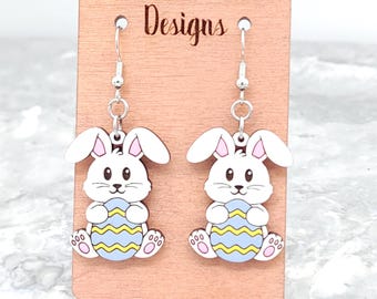 Può includere: Un paio di orecchini a forma di coniglio bianco che tengono uova di Pasqua a strisce blu e gialle. Gli orecchini sono su una carta espositiva in legno con il testo "Wilder Mind Designs".