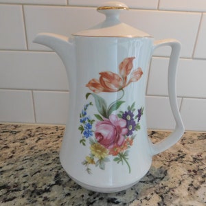 Puede incluir: Cafetera de porcelana blanca con un diseño floral con flores rosas, naranjas, amarillas, azules y moradas. La cafetera tiene un borde dorado alrededor de la tapa.