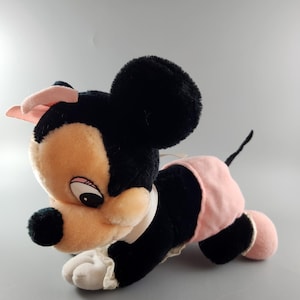 May include: A plush toy of a cartoon mouse, featuring black fur, a peach-colored face, and a pink bow. The mouse wears a pink skirt and has a white collar. The toy is lying down on a gray surface.