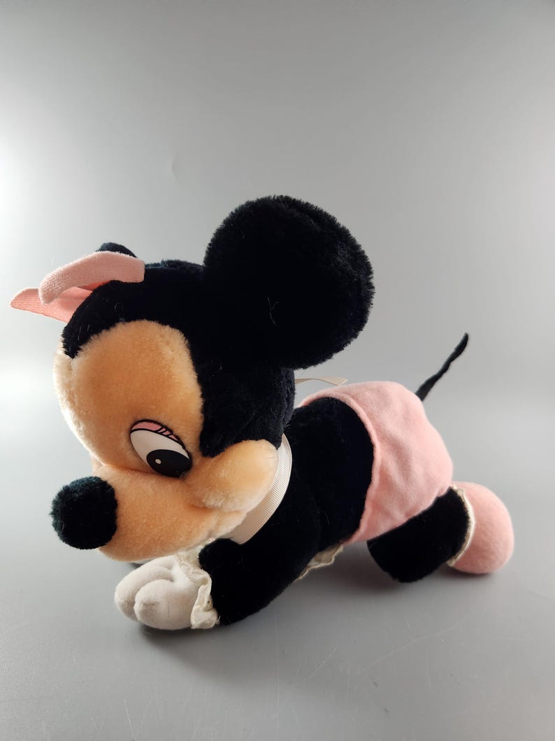 May include: A plush toy of a cartoon mouse, featuring black fur, a peach-colored face, and a pink bow. The mouse wears a pink skirt and has a white collar. The toy is lying down on a gray surface.