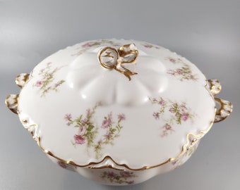 May include: White porcelain serving dish with a lid. The dish has a scalloped edge and is decorated with a floral pattern in shades of pink and green. The lid has a gold-coloured bow on top.
