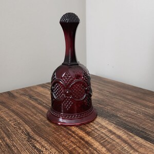 Puede incluir: Una campana de vidrio rojo oscuro con una superficie texturizada y un diseño decorativo. La campana tiene una parte superior redondeada y una base ancha.