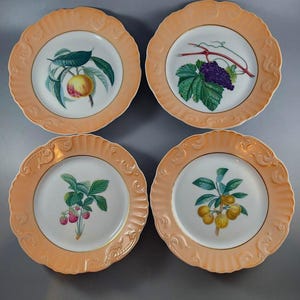 May include: Four decorative plates with peach-colored scalloped rims and gold trim. Each plate features a different fruit illustration: a peach, grapes, strawberries, and yellow fruit. The plates are arranged on a gray surface.