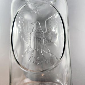 May include: Transparent glass jar with an embossed oval design featuring an eagle, star, and shield. The jar has a wide mouth and a circular base. The design is centered on the jar's body, and the glass is clear.