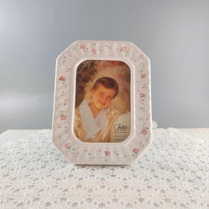 May include: A white ceramic photo frame with a floral design and a picture of a child. The frame is octagonal with a scalloped edge and small pink and green floral accents. The photo is 3.5 x 5 inches.