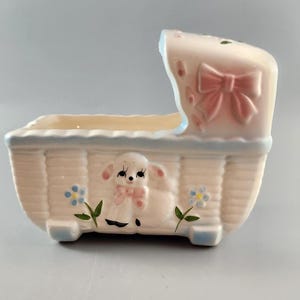 May include: A vintage cream ceramic baby cradle planter with blue detailing. The planter features a lamb with a pink bow, pink flowers, and a pink bow on the hood. A charming decorative piece.