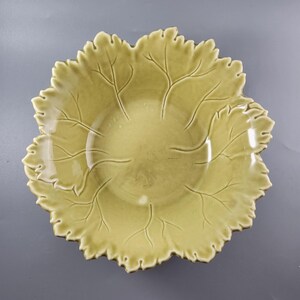 Puede incluir: Un cuenco de cerámica de color amarillo pálido con forma de hoja grande. El cuenco presenta un acabado brillante con detalles de nervaduras en relieve que irradian desde el centro hacia los bordes festoneados y serrados, con forma redonda.