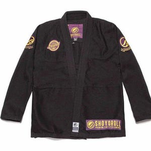 Puede incluir: Kimono de Jiu-Jitsu negro con parches bordados dorados y morados. El nombre de la marca "SHOYOROLL" es visible. El kimono tiene un cuello tradicional y costuras reforzadas. Ideal para entrenamiento.