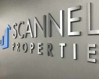 Könnte beinhalten: Ein silbernes Metallschild mit dem Text "SCANNELL PROPERTIES" in erhabenen Buchstaben. Das "S" in "SCANNELL" ist größer als die anderen Buchstaben und hat eine andere Silberfarbe. Links neben dem Text befindet sich eine blaue Grafik eines nach oben zeigenden Pfeils.
