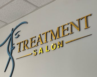 Könnte beinhalten: Wandmontiertes Schild für einen Salon mit goldfarbenen Spiegelbuchstaben, die "TREATMENT SALON" lauten. Das Schild ist an einer hellgrauen Wand angebracht, mit einem blauen und schwarzen Dekorationselement auf der linken Seite. Eine Überwachungskamera ist oben montiert.