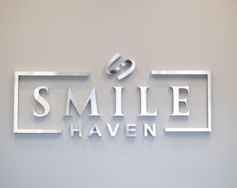 Könnte beinhalten: Ein silbernes Schild mit den Worten "SMILE HAVEN" in einem rechteckigen Rahmen. Das Wort "SMILE" ist in einer größeren Schriftart als das Wort "HAVEN".