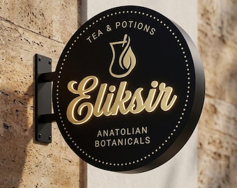 Könnte beinhalten: Rundes, schwarzes Schild mit goldfarbener Schrift, das "Tea & Potions", "Eliksir" und "Anatolian Botanicals" anzeigt. Es zeigt eine Teetasse-Grafik und ist an einer Wand befestigt. Das Schild ist an einer schwarzen Metallhalterung befestigt.