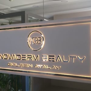 May include: A white sign with a gold border and a gold logo that says "NB" inside a circle. The sign also says "NOVADERM BEAUTY GÜLSEN ATALAY" in gold letters.