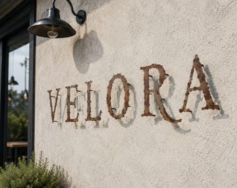 Könnte beinhalten: Eine strukturierte cremeweiße Wand zeigt das Wort VELORA in großen, rostigen Metallbuchstaben, die mit sichtbaren Befestigungen montiert sind. Eine schwarze, kuppelförmige Außenleuchte ist über dem Schriftzug angebracht und wirft einen Schatten.