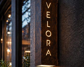 Könnte beinhalten: Ein rechteckiges, bronzefarbenes Schild mit dem Wort "VELORA" vertikal in beleuchteten weißen Buchstaben. Das Schild ist an einer dunklen, strukturierten Wand angebracht, mit warmer Beleuchtung von unten.