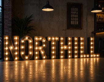 Könnte beinhalten: Große, beleuchtete Leuchtbuchstaben mit der Aufschrift "NORTHILL" mit warmen, leuchtenden Glühbirnen. Die Buchstaben bestehen aus verwittertem Metall und werfen ein warmes Licht auf den Boden. Ein Schild im Hintergrund lautet "GOOD PEOPLE DRINK GOOD BEER".