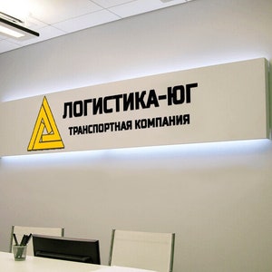 May include: A white sign with black text that reads "Логистика-лог Транспортная компания" and a yellow triangle logo.
