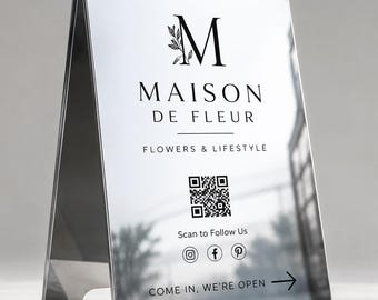 Könnte beinhalten: Ein silberfarbenes, reflektierendes A-Aufsteller-Schild mit dem schwarzen Text Maison de Fleur, Flowers & Lifestyle, einem großen M mit Blattmotiven, einem QR-Code, Social-Media-Symbolen sowie dem Text Scan to Follow Us und Come In, We're Open.