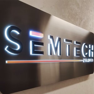 May include: A rectangular metal sign with the text "SEMTECH" in a modern font, illuminated with white light. The sign also includes the text "EST. 1968" and a horizontal orange and yellow light bar. The sign is mounted on a textured wall.
