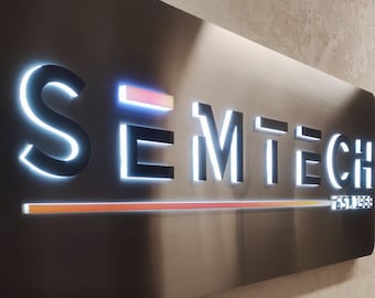 Könnte beinhalten: Rechteckiges Metallschild mit dem Text "SEMTECH" in einer modernen Schriftart, beleuchtet mit weißem Licht. Das Schild enthält auch den Text "EST. 1968" und einen horizontalen orange-gelben Lichtbalken. Das Schild ist an einer strukturierten Wand befestigt.