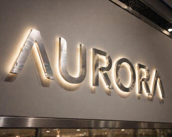 Könnte beinhalten: Das Wort "AURORA" in großen, gebürsteten Metallbuchstaben ist an einer hellen Wand angebracht. Jeder Buchstabe ist mit einem warmen, weichen Schein hinterleuchtet, was einen auffälligen Kontrast erzeugt. Das Schild befindet sich über einer reflektierenden Oberfläche.
