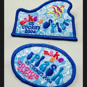 K&ouml;nnte beinhalten: Zwei gestickte Aufn&auml;her mit blauen R&auml;ndern. Einer hat eine einzigartige Form, der andere ist oval. Beide haben einen hellblauen Hintergrund mit den Worten "make a splash" und "GS Cookies 2004". Der einzigartige Aufn&auml;her hat ein farbenfrohes Fischdesign.