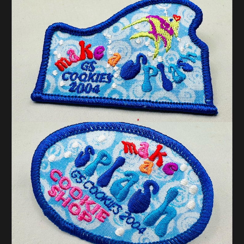 K&ouml;nnte beinhalten: Zwei gestickte Aufn&auml;her mit blauen R&auml;ndern. Einer hat eine einzigartige Form, der andere ist oval. Beide haben einen hellblauen Hintergrund mit den Worten "make a splash" und "GS Cookies 2004". Der einzigartige Aufn&auml;her hat ein farbenfrohes Fischdesign.