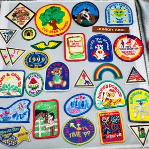 May include: A collection of colorful Girl Scout patches with various designs and text. Patches include images of trees, animals, rainbows, and text such as "Share the Season" and "Cookie Volunteer".