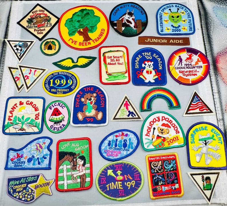 K&ouml;nnte beinhalten: Eine Sammlung farbenfroher Pfadfinderinnen-Abzeichen mit verschiedenen Designs und Texten. Die Abzeichen zeigen Bilder von B&auml;umen, Tieren, Regenb&ouml;gen und Texte wie "Share the Season" und "Cookie Volunteer".
