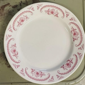 Può includere: Piatto in ceramica bianca con un motivo floreale rosa sul bordo. Il design include fiori stilizzati e bordi smerlati. Un design classico del piatto, adatto all'uso quotidiano.