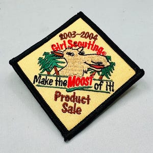 K&ouml;nnte beinhalten: Ein quadratischer Pfadfinderinnen-Aufn&auml;her mit beigem Hintergrund und schwarzem Rand. Der Aufn&auml;her zeigt eine Elch-Grafik mit dem Text "2003-2004 Girl Scouting", "Make the MOOST of HI" und "Product Sale".