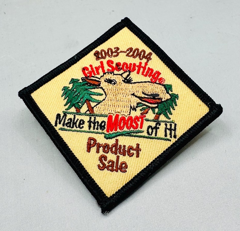 K&ouml;nnte beinhalten: Ein quadratischer Pfadfinderinnen-Aufn&auml;her mit beigem Hintergrund und schwarzem Rand. Der Aufn&auml;her zeigt eine Elch-Grafik mit dem Text "2003-2004 Girl Scouting", "Make the MOOST of HI" und "Product Sale".