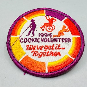 K&ouml;nnte beinhalten: Runder, gestickter Aufn&auml;her mit lila Rand und wei&szlig;er Mitte. Der Aufn&auml;her zeigt den Text "1994 Cookie Volunteer" und "We've got it... Together." Silhouetten von Personen sind in Orange und Lila dargestellt, mit einem Farbverlauf von Orange, Gelb und Lila.